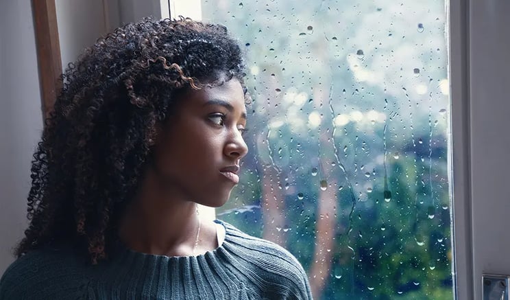 A young woman looks worried or sad, staring out of a rainy window pane