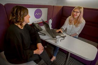 Two women sat laughing at a desk together. They are sat within a booth with purple seats.