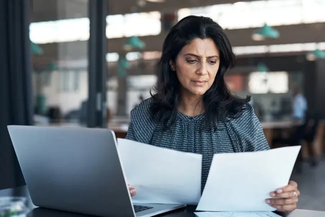 A woman in an office environment is sat at her laptop, looking at two pieces of paper in her hands, looking concerned. 