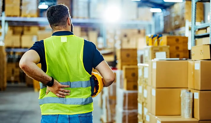 A man in a high-vis jacket faces away from the camera, supporting his back with one hand in a warehouse surrounded by boxes