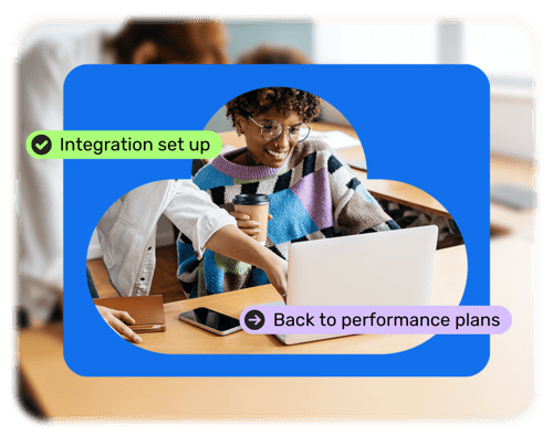 A person holding a coffee is sat at a desk looking at their laptop. Another person is pointing at the screen. It displays a notification in a green bubble that reads 'Integration set up'. There's another purple bubble that reads 'Back to performance plans'. 