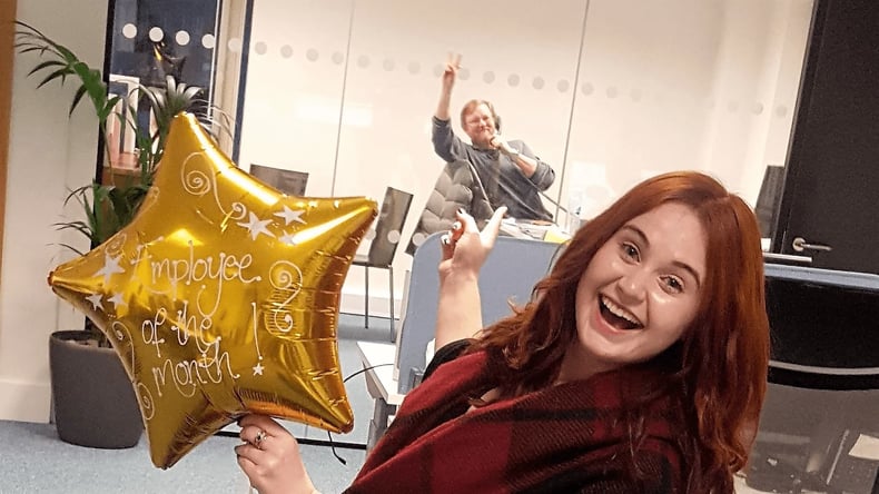 A woman is smiling and holding a star shaped golden "employee of the month" balloon. She is pointing behind her at her colleague who is on the phone in a meeting room.