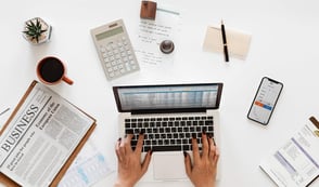 A person is working on their laptop whilst sat at their desk. The laptop is surrounded by a newspaper, coffee cup, succulent, calculator, notepad and a phone.