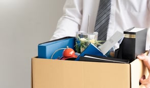 A man in a white shirt and striped tie is carrying a cardboard box full of his office supplies.
