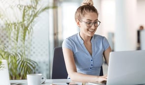 A woman in a blue shirt is working at a desk on her laptop. She has a white mug on her table and has a palm plant behind her.