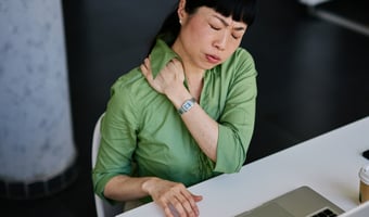 A female employee wearing glasses looks stressed while sitting at her desk, surrounded by paperwork and holding her head in her hands. Behind her, an out-of-focus colleague works at another desk. The scene reflects the pressure and confusion that can come from overlooked health and safety responsibilities in a growing business.