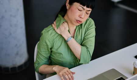 A female employee wearing glasses looks stressed while sitting at her desk, surrounded by paperwork and holding her head in her hands. Behind her, an out-of-focus colleague works at another desk. The scene reflects the pressure and confusion that can come from overlooked health and safety responsibilities in a growing business.