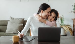 Parent working from home on a laptop while hugging their child, illustrating flexible working for parents and carers in SMEs.