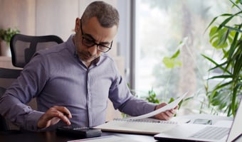 A middle-aged employer with short grey-flecked hair and black-rimmed glasses reviews National Minimum Wage costs, using a calculator and documents at a home office desk, with a laptop open beside him