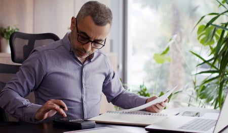 A middle-aged employer with short grey-flecked hair and black-rimmed glasses reviews National Minimum Wage costs, using a calculator and documents at a home office desk, with a laptop open beside him