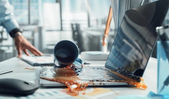 Spilled coffee pouring from an overturned mug across a laptop keyboard and desk in a modern office.