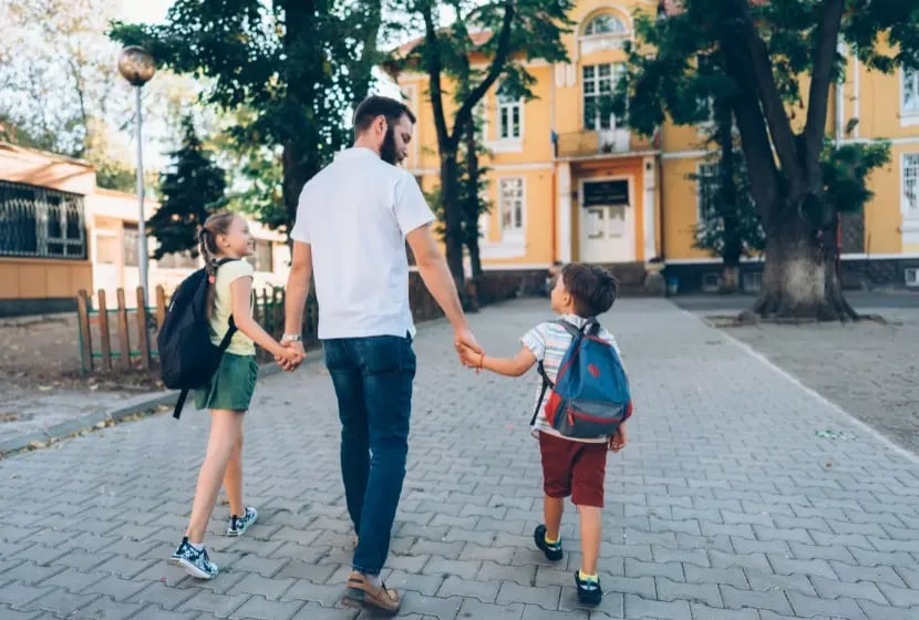A view from behind shows a bearded man in a white shirt and jeans holding hands with a girl in pigtails and a boy with backpacks as they walk on a grey stone path away from a large yellow building with many windows and trees around it