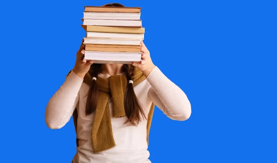 A person with long brown hair in pigtails holds a tall stack of books in front of their face against a solid blue background. The person is wearing a white long-sleeved shirt and has a brown sweater draped over their shoulders. The stack of books has varying thicknesses and shades of brown and white on their spines and pages. The person's hands are visible holding the books up.