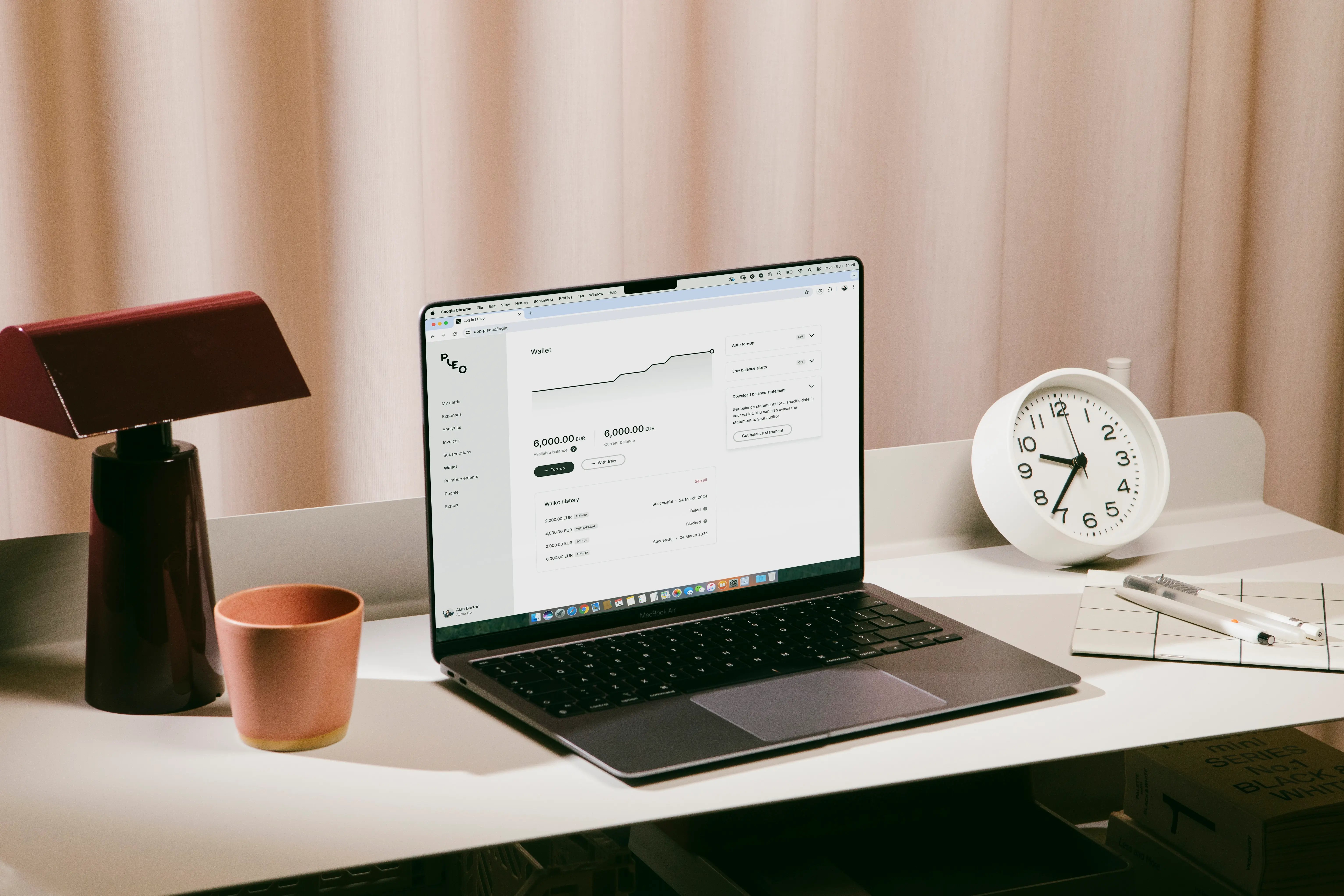 A laptop displaying the Pleo business expense management platform, showing a graph and financial figures, in a softly lit workspace with a lamp, drink, and clock.