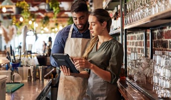 Two hospitality staff members review a tablet at a bar while managing staggered shift patterns.