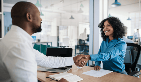 a man and a woman sit across an office table from each other, shaking hands. The woman is facing the camera and is smiling