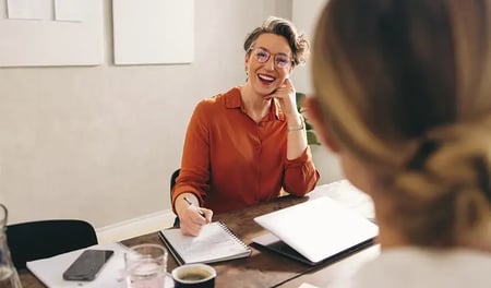 Businesswoman in a meeting, smiling and taking notes at a desk with a laptop.
