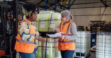 Two employees in high-visibility vests review paperwork together in a warehouse, focusing on workplace health and safety.