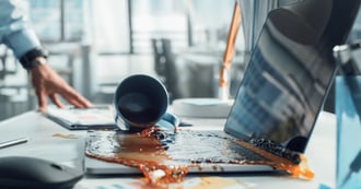 Spilled coffee pouring from an overturned mug across a laptop keyboard and desk in a modern office.