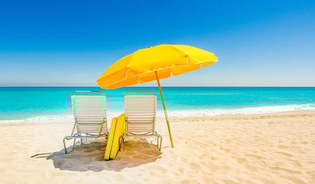 Two deck chairs and a yellow parasol are on a white sandy beach, near an aqua blue sea.