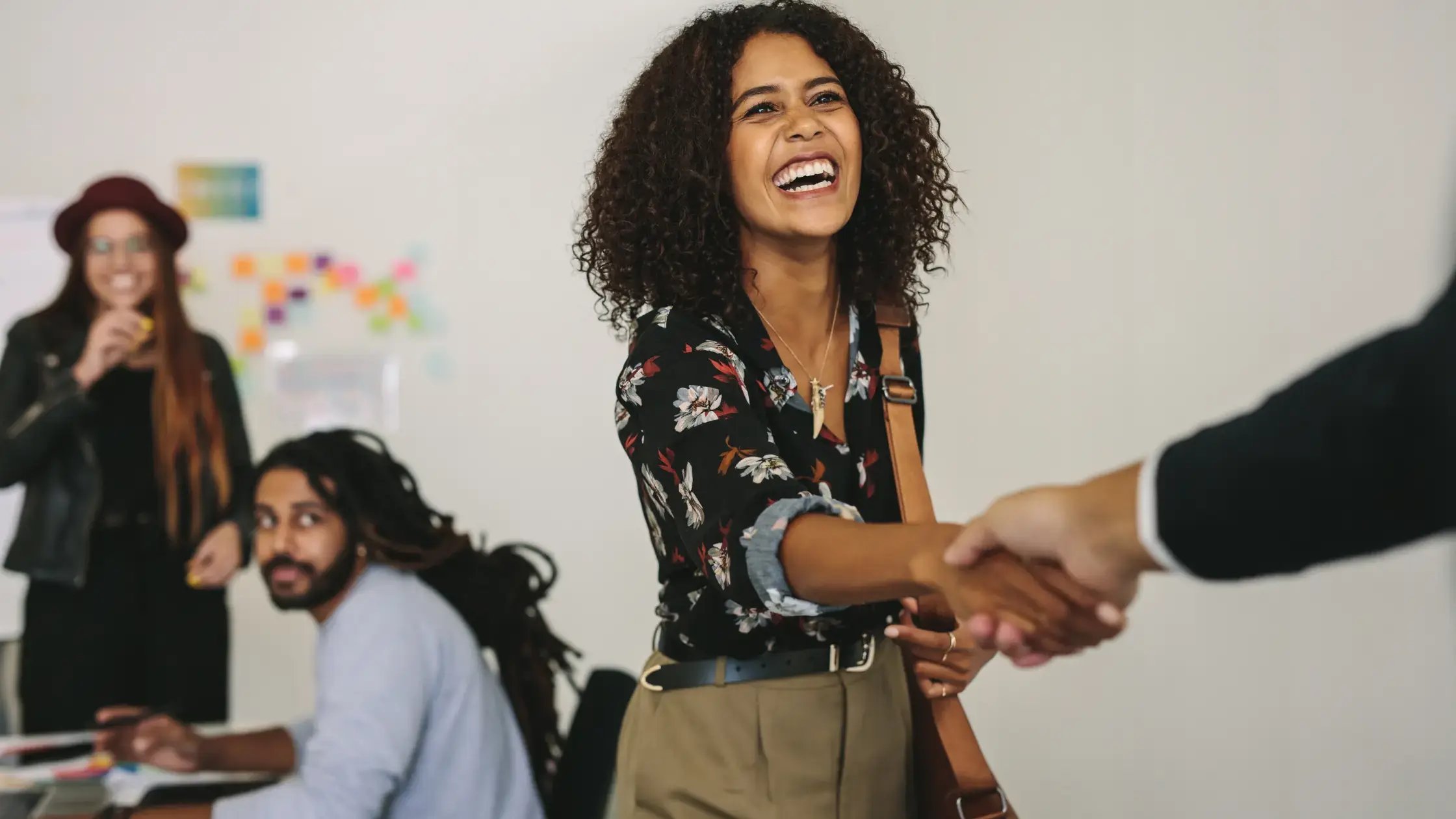 In an office environment, a person with dark curly hair, wearing a patterned shirt and tan trousers, beams while shaking hands with someone whose arm enters the frame from the right. Two other people are visible in the background near a whiteboard.