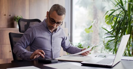 A middle-aged employer with short grey-flecked hair and black-rimmed glasses reviews National Minimum Wage costs, using a calculator and documents at a home office desk, with a laptop open beside him