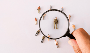 A magnifying glass is hovering over a miniature toy model of a man in a suit. There are several other miniature models of people surrounding him but they are not enlarged by the magnifying glass. They are placed on a white background.