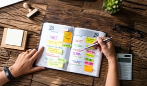 A person writing in a planner filled with colorful sticky notes and tasks. The wooden desk holds a calculator, glasses, a plant, and a notepad. The planner shows a busy two-day schedule of work and personal tasks.