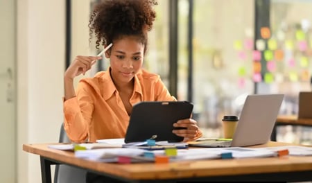 Businesswoman in an orange shirt working at a desk, holding a tablet and pen while reviewing documents with a laptop and office supplies nearby.