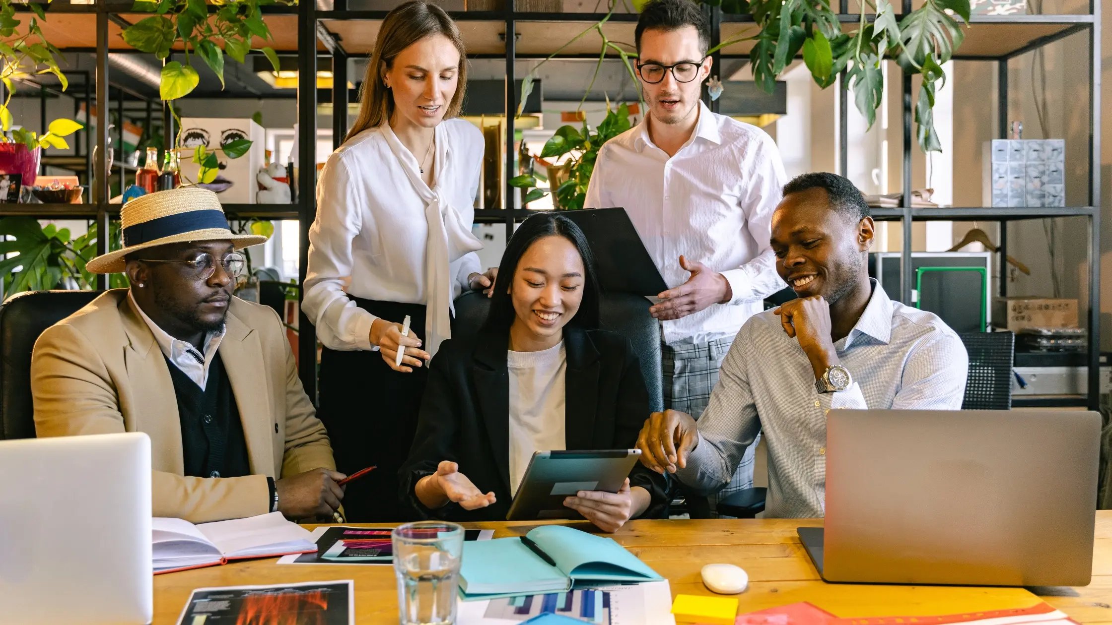 A group of five professionals collaborate in a modern office space with shelving and plants in the background. Three people are seated at a wooden desk with laptops and documents, while two others stand behind them, one holding a clipboard/tablet. They appear to be discussing the information displayed.