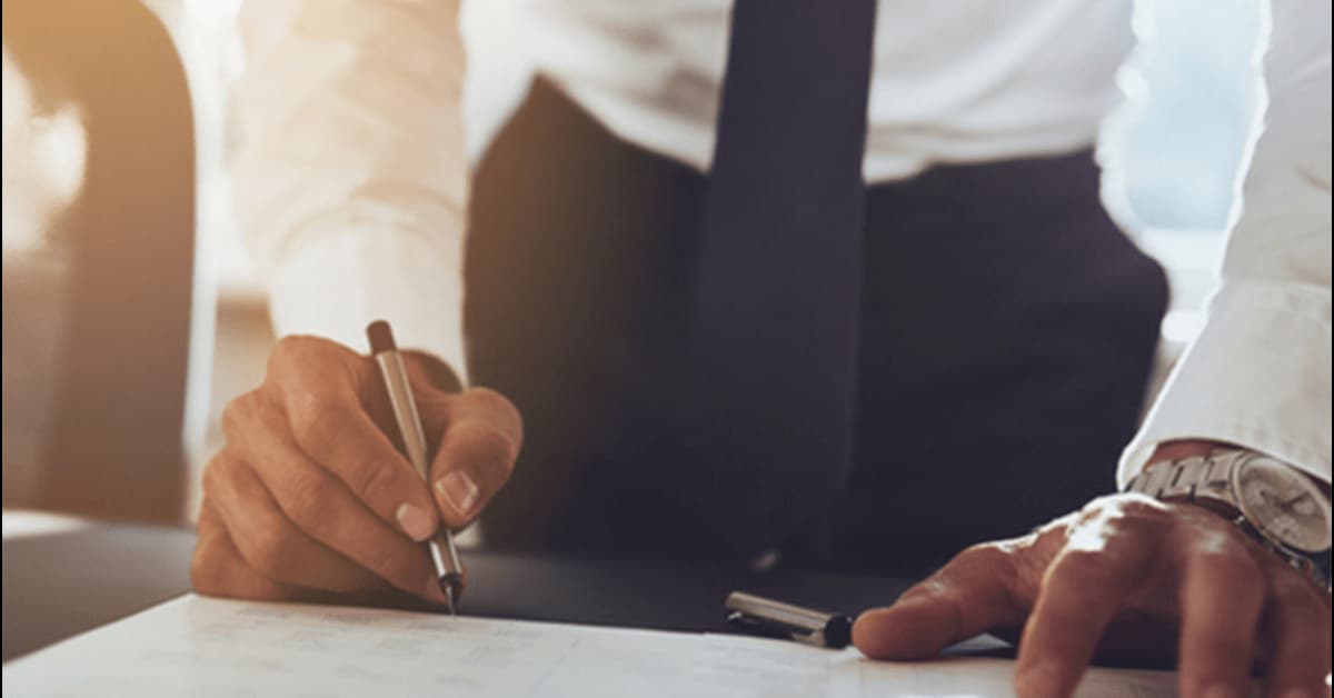 A man wearing a suit is signing some paperwork with a silver pen.