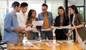 A group of employees stand around a table, reviewing documents. They seem to be presenting them to a man in the centre, who appears to be the manager.