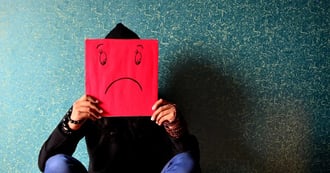 A person is sat on the floor against a blue wall. They are holding a bright red piece of paper over their face which has a sad face on it.