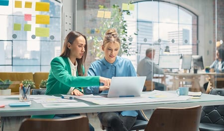 Two women colleagues sit in a modern office with glass walls. One is pointing at the other