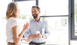 A man and a woman hold coffees in an office environment. They look happy and are smiling. 