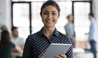 Smiling woman in a navy polka-dot shirt holding a tablet, standing in a bright office with blurred colleagues in the background.