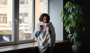 A woman stands next to a window and a plant wrapped in a big jumper holding a mug and also her phone, which she's looking at. 