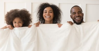 A woman, a man and their daughter holding a duvet up to necks and smiling. The parents are having duvet days given to them by their work. A duvet day is a day that an organisation agrees employees can take off without advance notice.