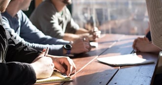 Four colleagues are sat around a wooden table together. They are having a meeting and writing things down in their notebooks.