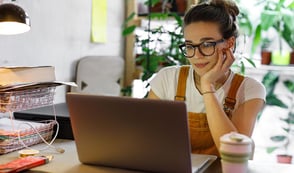 A woman wearing a white shirt and yellow dungarees is working from home under flexible working arrangements. The woman is sat at her desk with a laptop in front of her. Her laptop is surrounded by paperwork, her phone, a cable and a coffee cup. Behind her is a shelf full of plants.