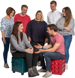 A team of seven people laughing together. Two of the colleagues are sat on cushioned stools whilst the rest of the team surround them.