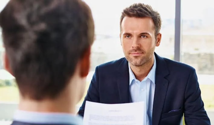 Two people are sat at a desk opposite each other. One man, wearing a suit, is facing the other and holding a piece of paper in his hand.