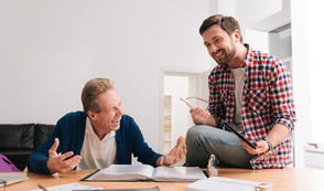 Two men are in conversation in an office environment. Their body language is relaxed and they're laughing. 