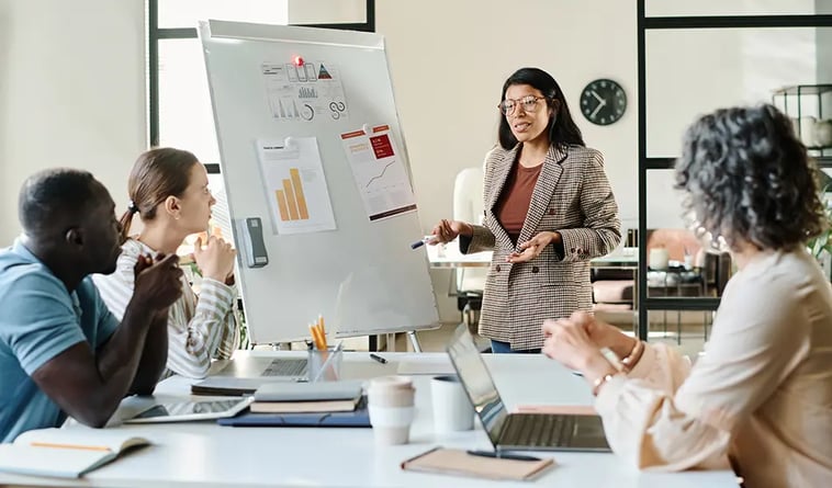 A woman stands in front of a whiteboard in a team meeting