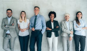 A diverse group of 6 employees of different ages, genders and ethnicities stand against a wall in a line, smiling. 