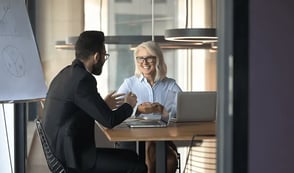 A man and a woman are sitting at a wooden desk with laptops and paperwork in front of them. They are having a meeting in one of their office rooms, smiling and chatting to each other. 