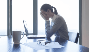 A woman is sat at a wooden desk with her laptop in front of her, surrounded by paperwork. She has her head in her hands and is looking very stressed. Her mental health in the workplace is declining.