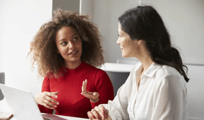 Two female colleagues are sat down together and having a one to one meeting. One woman is wearing a bright red jumper. She is talking to her colleague who is wearing a white blouse and has a laptop in front of her.