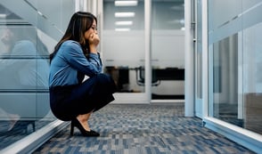 A woman is crouched down with her back against a glass office wall, with her chin in her hands. Her eyes are closed in an exasperated expression. This & her body language seem to say 
