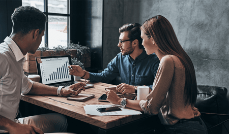 Two men and a woman are in an office, sat around a table, looking away from the camera and at a graph on a laptop. 